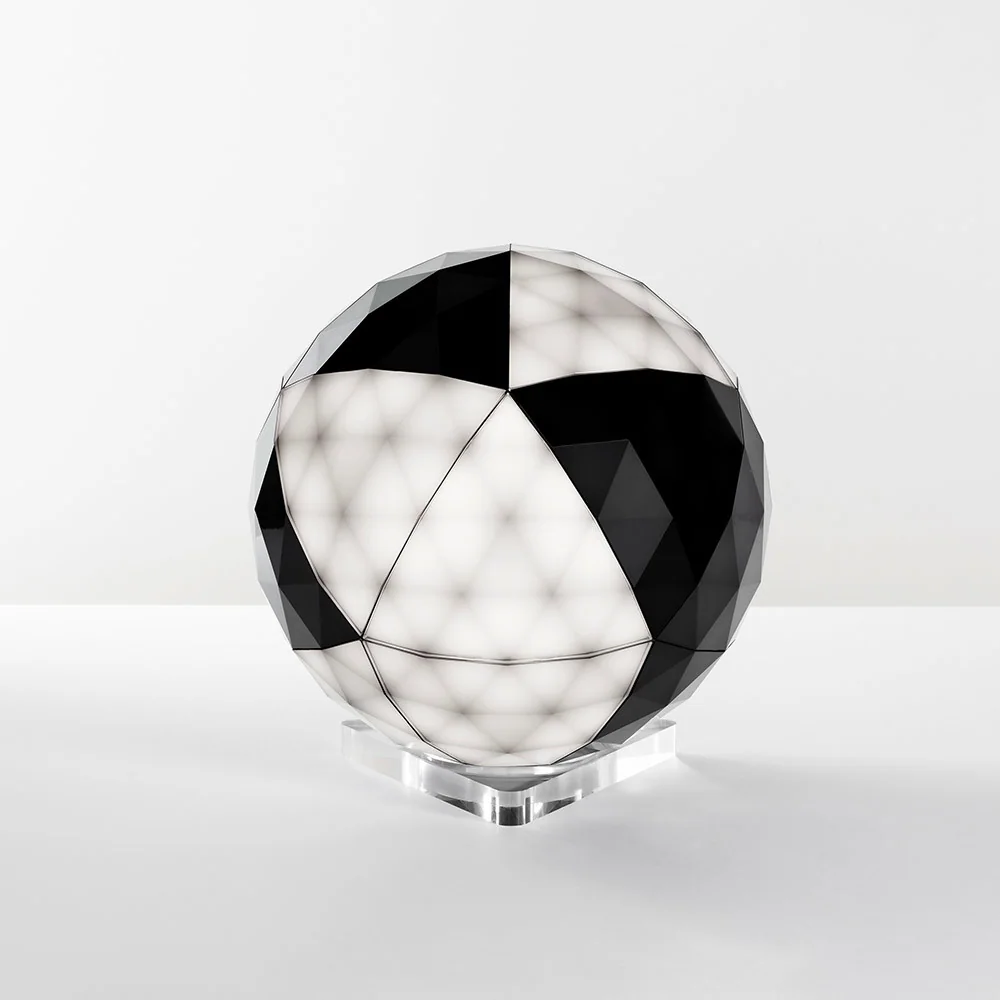 Artemide: Kugelförmiges Lichtobjekt mit abwechselnd dunklen und hellen polygonalen Flächen auf einem transparent wirkenden Sockel.