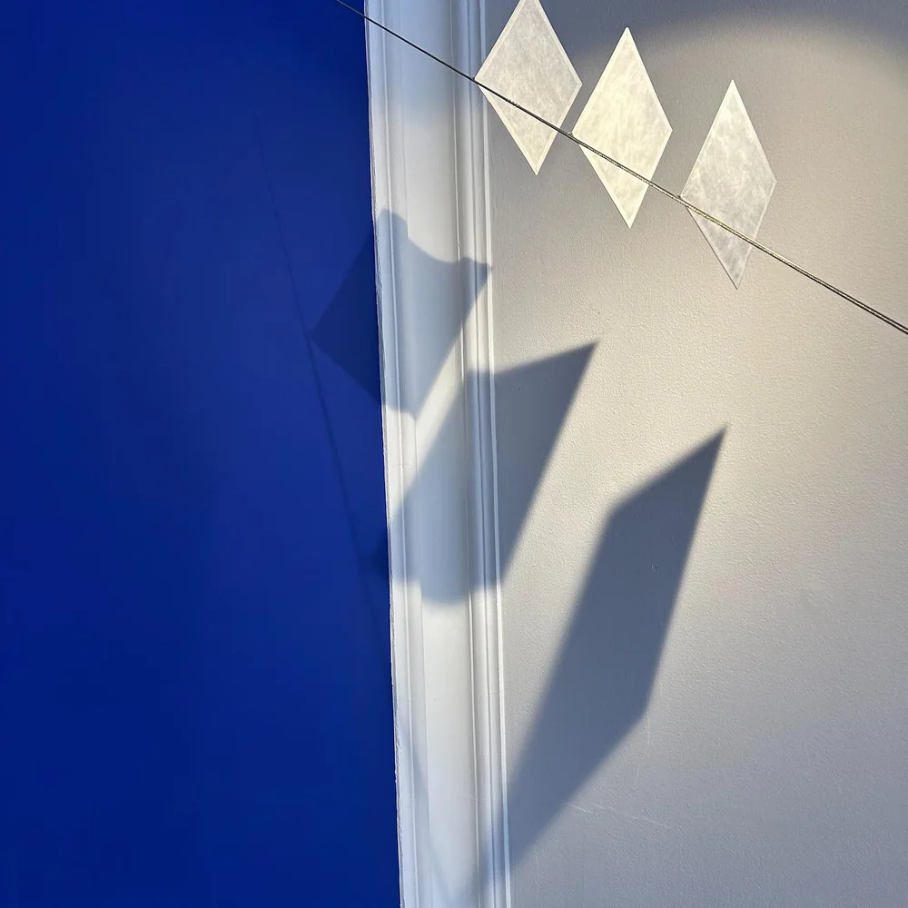Ingo Maurer: Geometrische helle Licht- und Schattenformen an einer blauen Wand als Detail einer Installation.