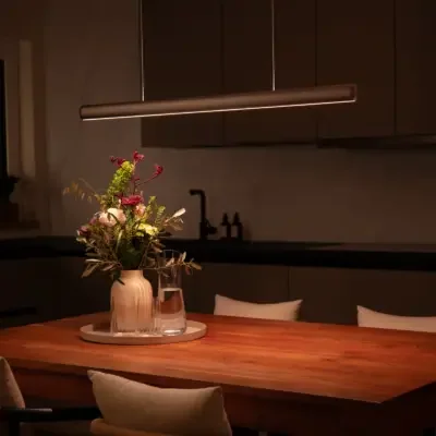 VILOOM: Bronzefarbene lineare Pendelleuchte über einem gedeckten Holztisch mit Blumen.