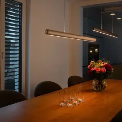 VILOOM: Lineare Pendelleuchte über einem Esstisch neben großen Fenstern in einem dunkleren Wohnraum.